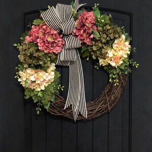 Pu&ograve; includere: Una ghirlanda realizzata con vite con ortensie rosa e crema e verde. Un nastro a righe bianche e nere &egrave; legato in un fiocco al centro della ghirlanda.