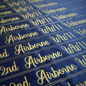 Puede incluir: Una pancarta de tela azul con letras doradas que dicen "82nd Airborne WWII Veterans". La pancarta se repite varias veces, creando un patr&oacute;n.