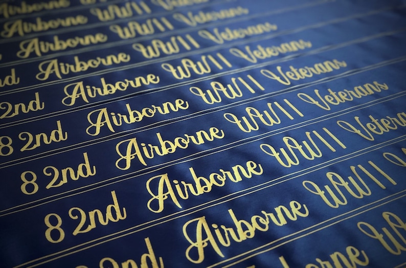 Puede incluir: Una pancarta de tela azul con letras doradas que dicen "82nd Airborne WWII Veterans". La pancarta se repite varias veces, creando un patr&oacute;n.