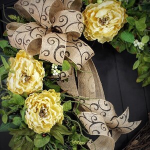 Pode incluir: Uma coroa de juta com um grande la&ccedil;o marrom e flores amarelas. O la&ccedil;o tem um padr&atilde;o de espiral marrom. A coroa &eacute; decorada com folhas verdes e bagas brancas.