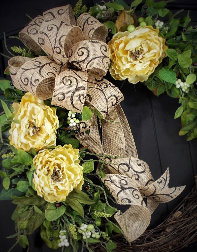 Pode incluir: Uma coroa de juta com um grande la&ccedil;o marrom e flores amarelas. O la&ccedil;o tem um padr&atilde;o de espiral marrom. A coroa &eacute; decorada com folhas verdes e bagas brancas.