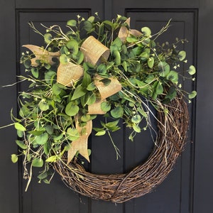 May include: A green eucalyptus wreath with a burlap bow. The wreath is made of natural twigs and is decorated with small white flowers.