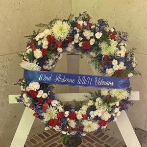 Puede incluir: Una corona azul y blanca con flores rojas y una cinta azul que dice "82nd Airborne WWII Veterans".