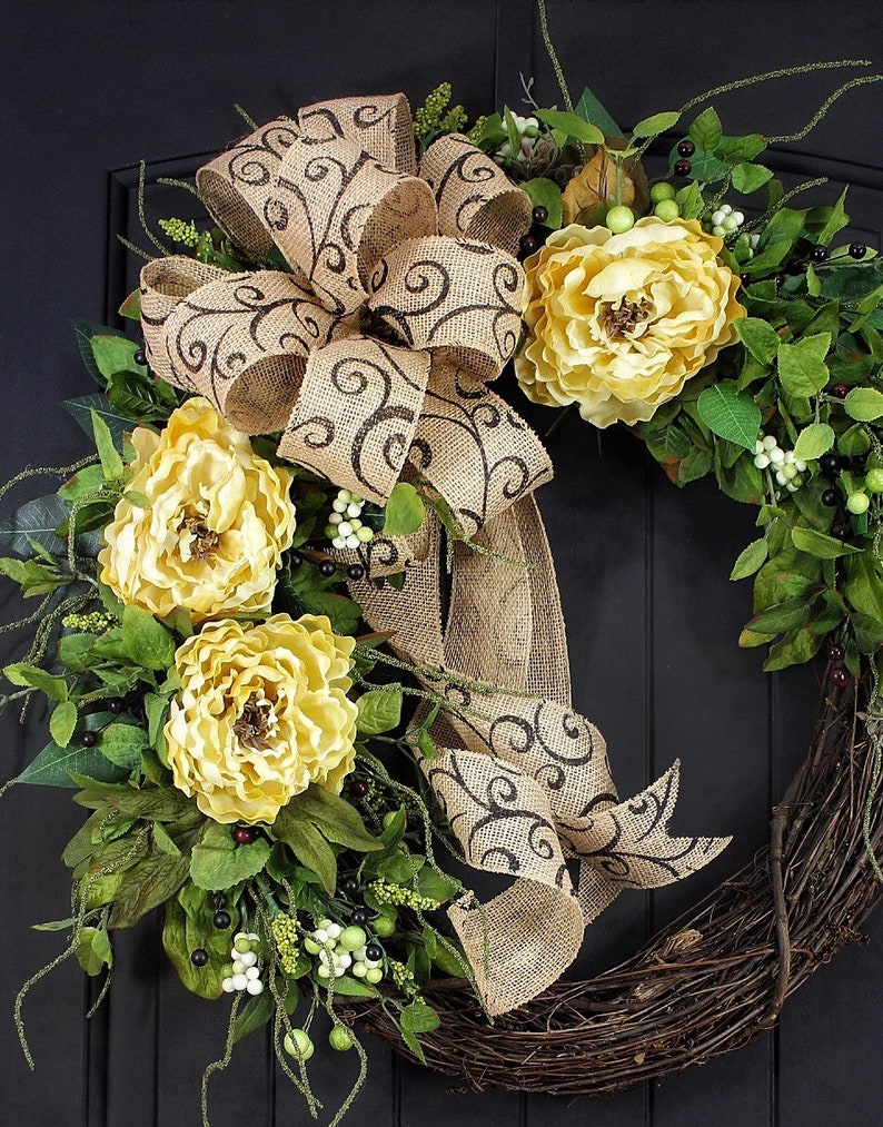 Pode incluir: Uma coroa de juta marrom com um grande la&ccedil;o e flores amarelas. O la&ccedil;o tem um padr&atilde;o de espiral marrom. A coroa &eacute; decorada com folhas verdes e bagas brancas.