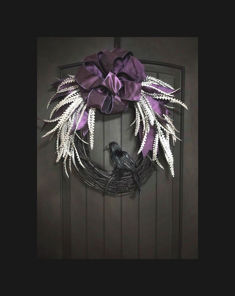 Puede incluir: Un cuervo negro posado en una corona de ramas con un lazo de sat&eacute;n morado y detalles de plumas blancas. La corona est&aacute; colgada en una puerta de madera oscura.