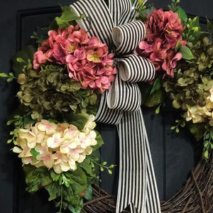 Pu&ograve; includere: Una ghirlanda di vite con fiori di ortensia rosa e crema e verde. Un nastro a righe bianche e nere &egrave; legato in un fiocco al centro della ghirlanda.