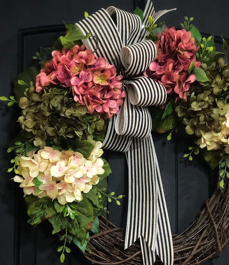 Pu&ograve; includere: Una ghirlanda di vite con fiori di ortensia rosa e crema e verde. Un nastro a righe bianche e nere &egrave; legato in un fiocco al centro della ghirlanda.