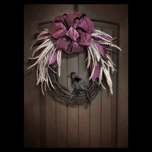 May include: A purple and white Halloween wreath with a black raven perched on a nest. The wreath is decorated with a large purple bow and white feathers.