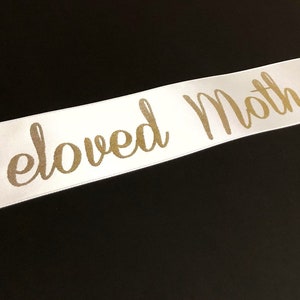 Puede incluir: Cinta de sat&eacute;n blanca con las palabras "Beloved Mother" impresas en letras doradas.