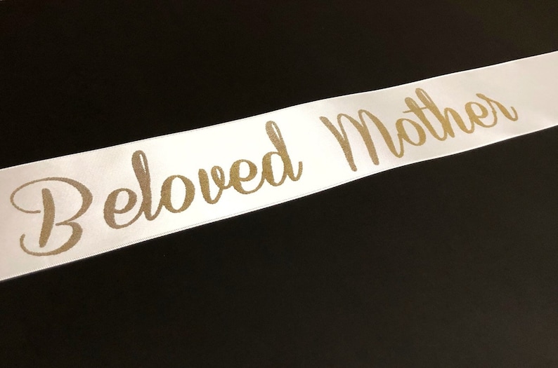 Puede incluir: Cinta de sat&eacute;n blanca con las palabras "Beloved Mother" impresas en letras doradas.