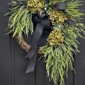 May include: A green and gold wreath with a black ribbon bow. The wreath is made of greenery and has a variety of green and gold flowers.