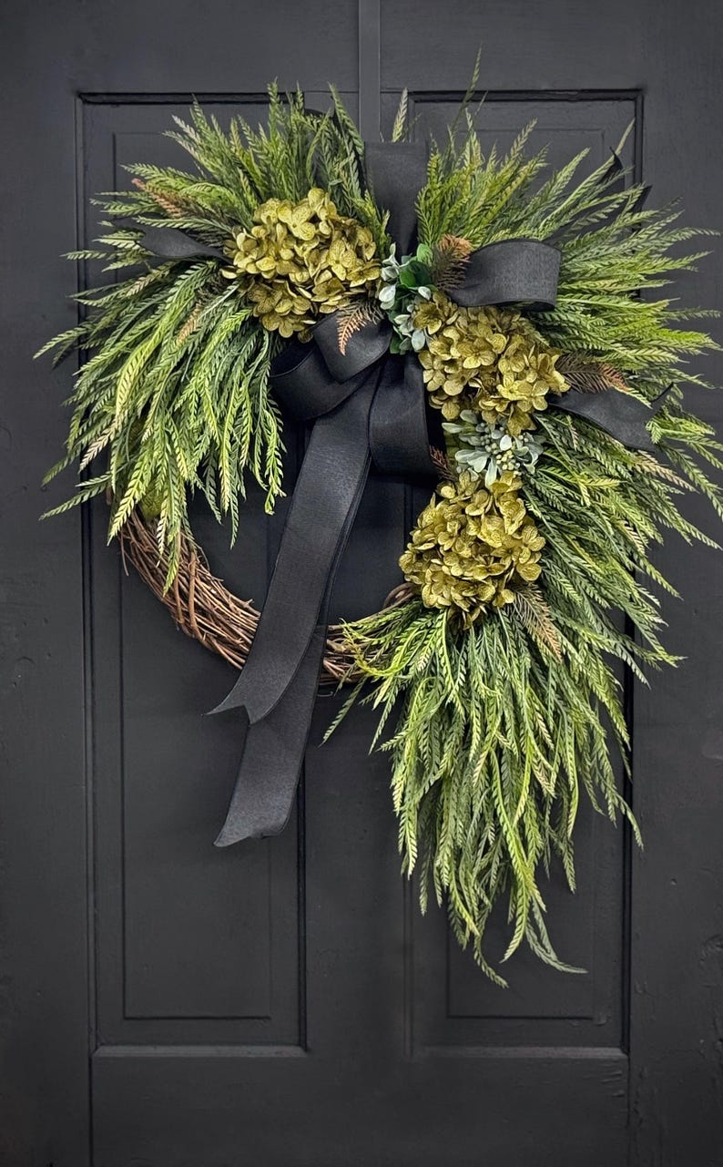 May include: A green and gold wreath with a black ribbon bow. The wreath is made of greenery and has a variety of green and gold flowers.