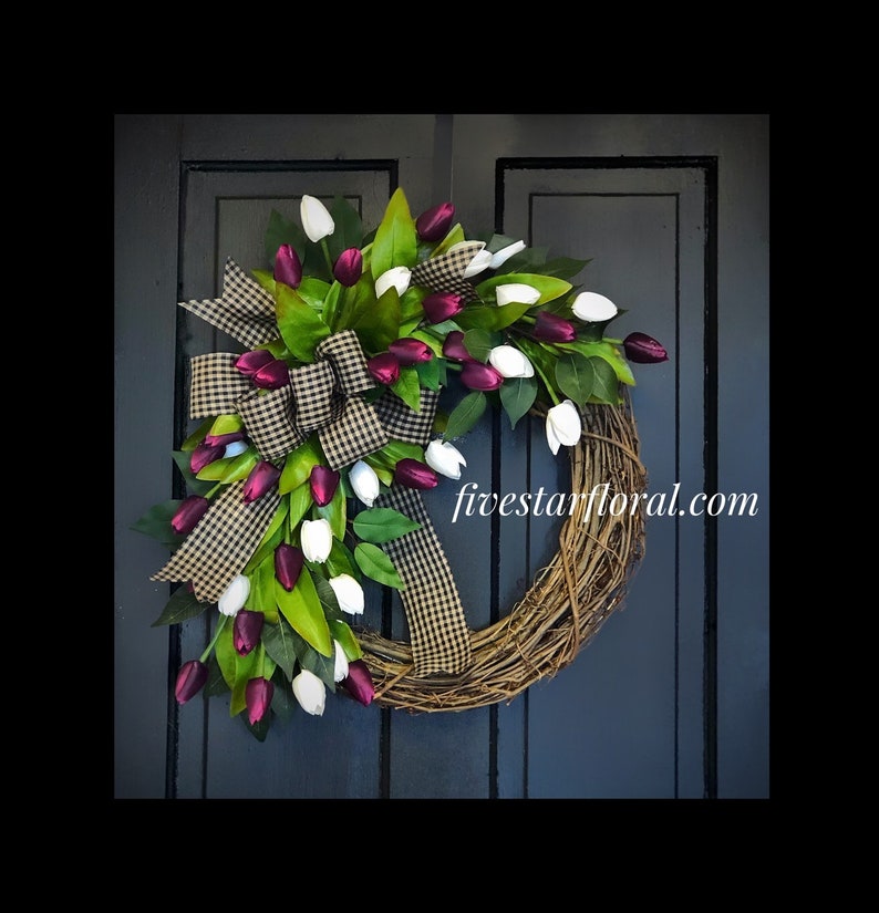 May include: A grapevine wreath decorated with white and burgundy tulips and a black and white checkered bow. The wreath is accented with green leaves and the text "fivestarfloral.com" is visible in the bottom right corner.