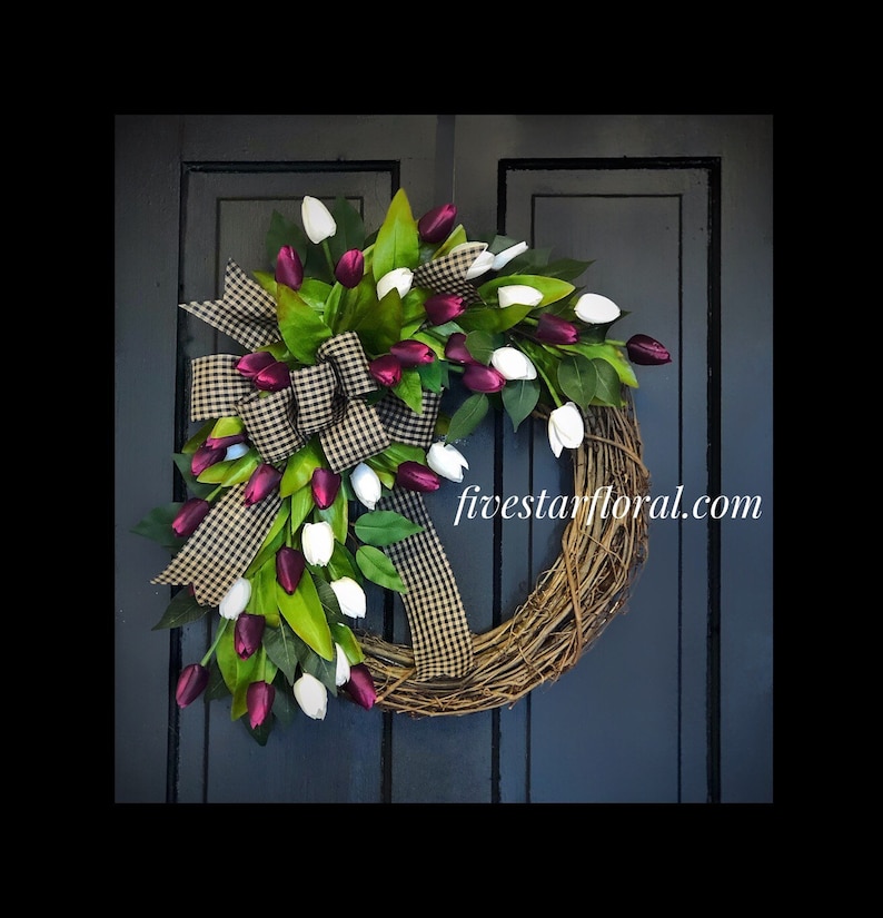 May include: A wreath made of grapevine with a black and white checkered ribbon and burgundy and white tulips. The wreath is decorated with green leaves and the text "fivestarfloral.com" is visible on the wreath.