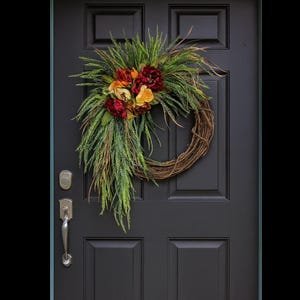 Puede incluir: Una corona decorativa con base de vid, adornada con un racimo de flores rojas, naranjas y amarillas, y acentuada con follaje verde. La corona cuelga de una puerta negra con manija y cerradura plateadas.