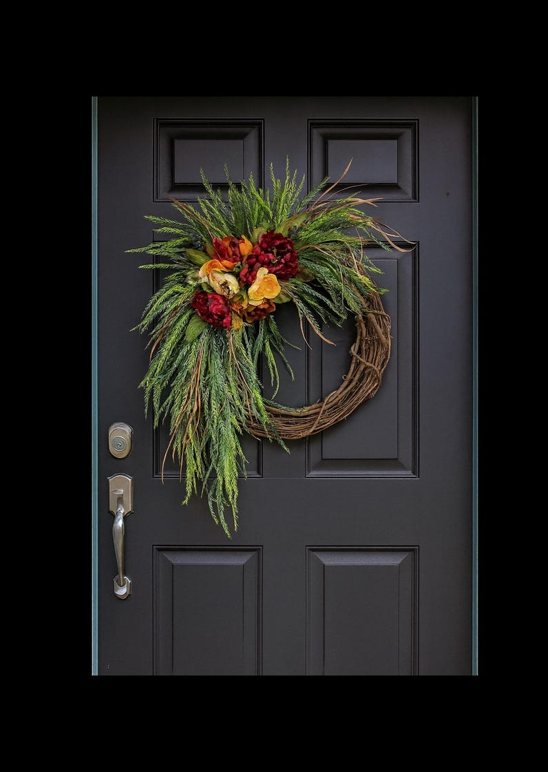 Puede incluir: Una corona decorativa con base de vid, adornada con un racimo de flores rojas, naranjas y amarillas, y acentuada con follaje verde. La corona cuelga de una puerta negra con manija y cerradura plateadas.