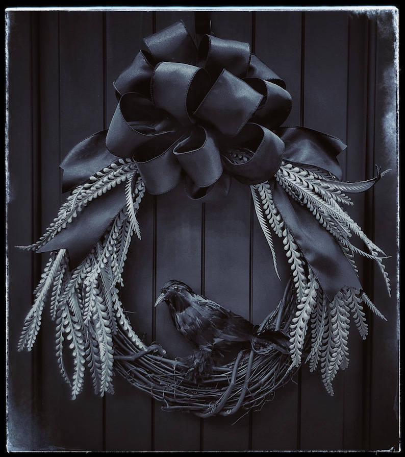 May include: A black crow sits in a wreath made of gray branches and black ribbon. The wreath is decorated with a large black bow.
