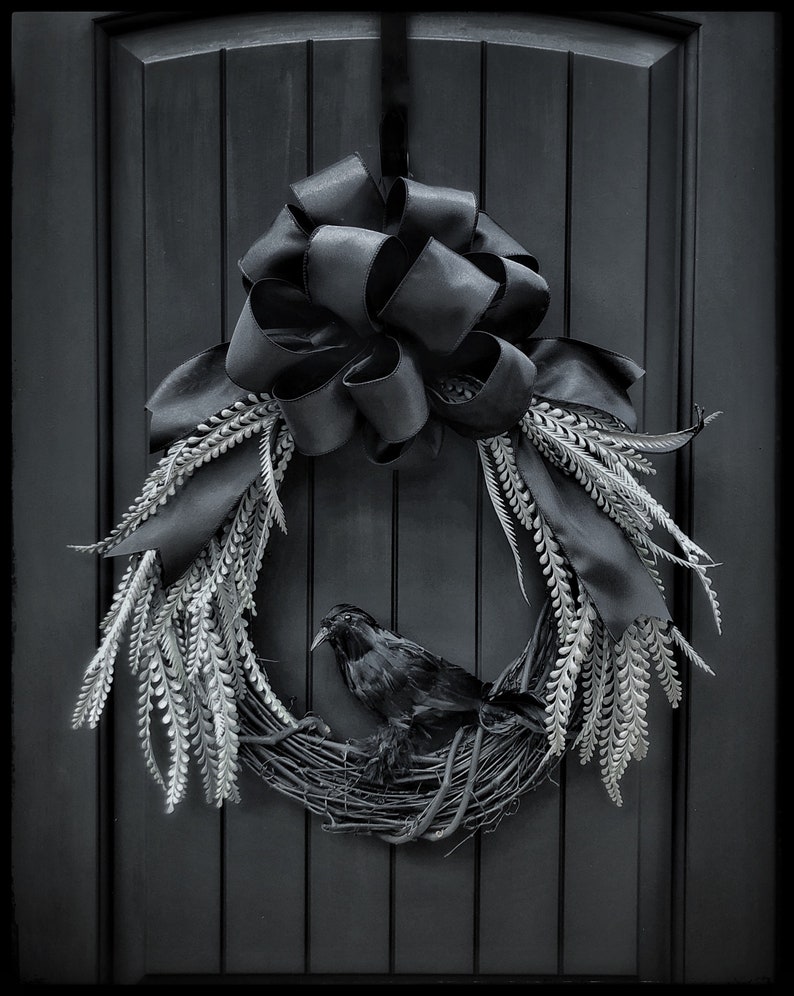 May include: A black raven sits in a black and white wreath with a black satin bow. The wreath is hanging on a black door.