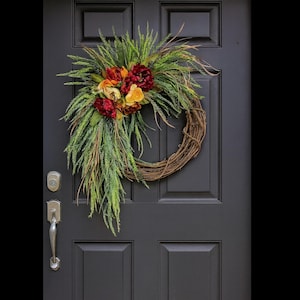 Puede incluir: Una corona r&uacute;stica de vid adornada con flores artificiales en tonos rojos, naranjas y amarillos, complementada con un exuberante follaje verde. La corona cuelga de una puerta gris oscura.