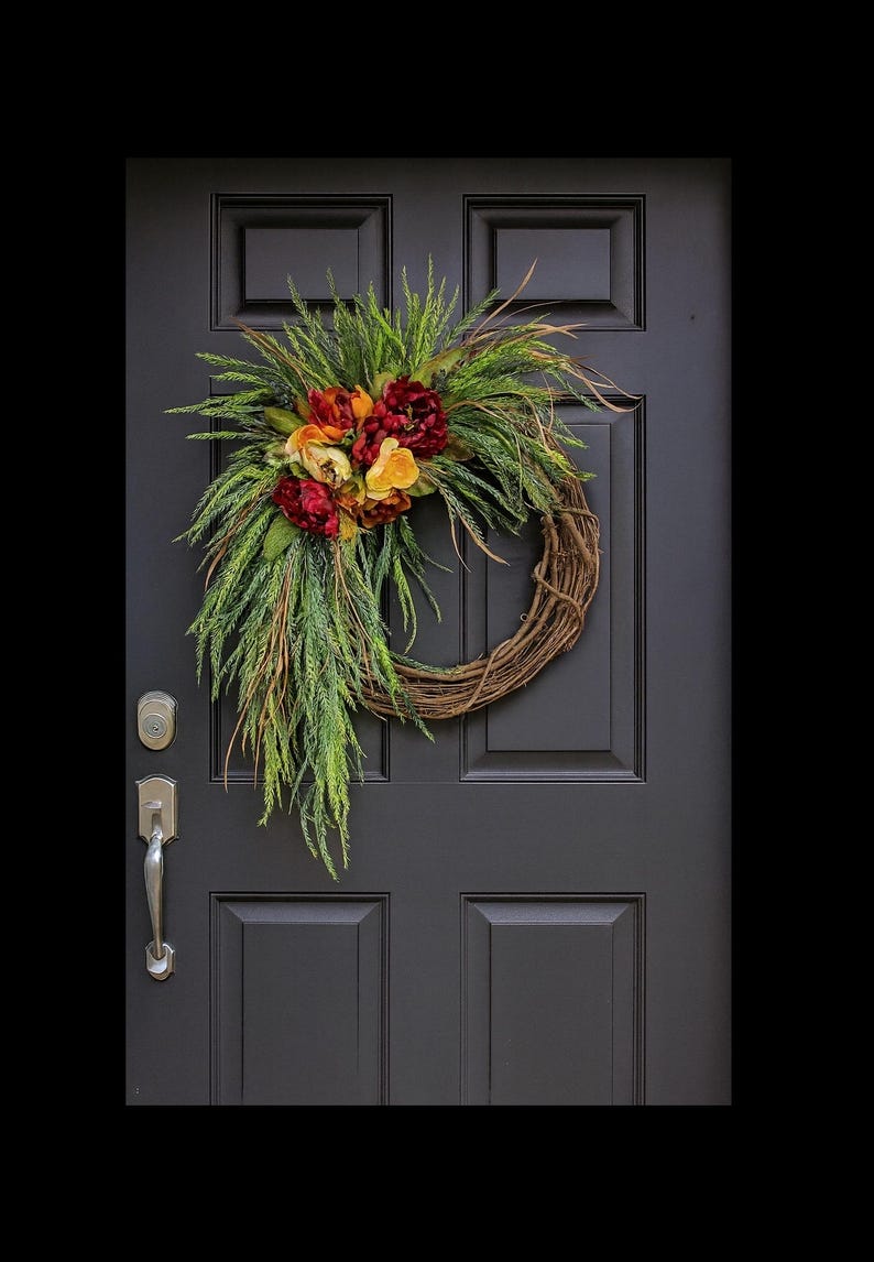 Puede incluir: Una corona r&uacute;stica de vid adornada con flores artificiales en tonos rojos, naranjas y amarillos, complementada con un exuberante follaje verde. La corona cuelga de una puerta gris oscura.