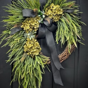 May include: A green and gold wreath with a black ribbon. The wreath is made of greenery and has a few clusters of gold flowers. The wreath is hanging on a black door.
