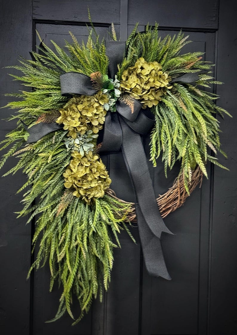 May include: A green and gold wreath with a black ribbon. The wreath is made of greenery and has a few clusters of gold flowers. The wreath is hanging on a black door.