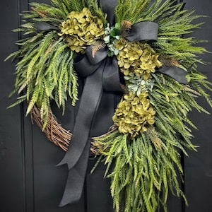 May include: A green and black wreath with a black ribbon bow. The wreath is made of greenery and has three clusters of green flowers.