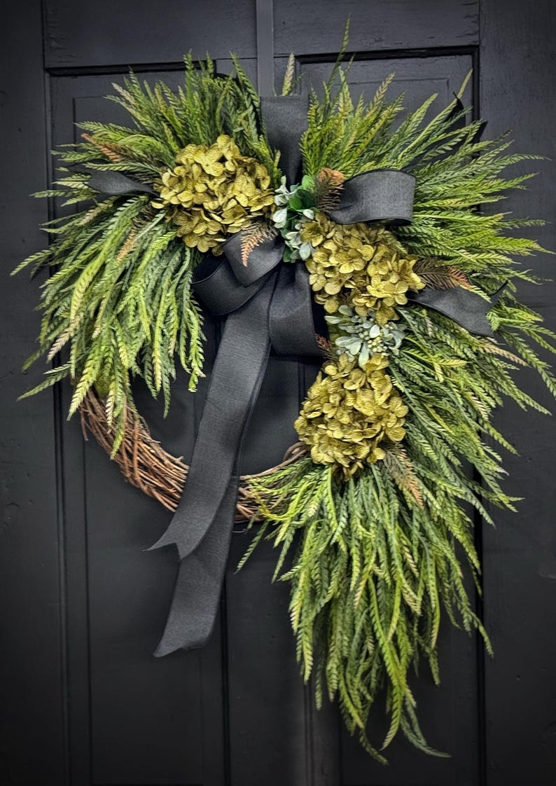 May include: A green and black wreath with a black ribbon bow. The wreath is made of greenery and has three clusters of green flowers.