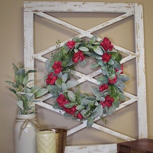 May include: A decorative white wooden wall hanging with a distressed finish, featuring a diamond-shaped lattice design. A wreath of red flowers and green leaves is centered within the lattice. A white jar with greenery, a candle, and books complete the display.