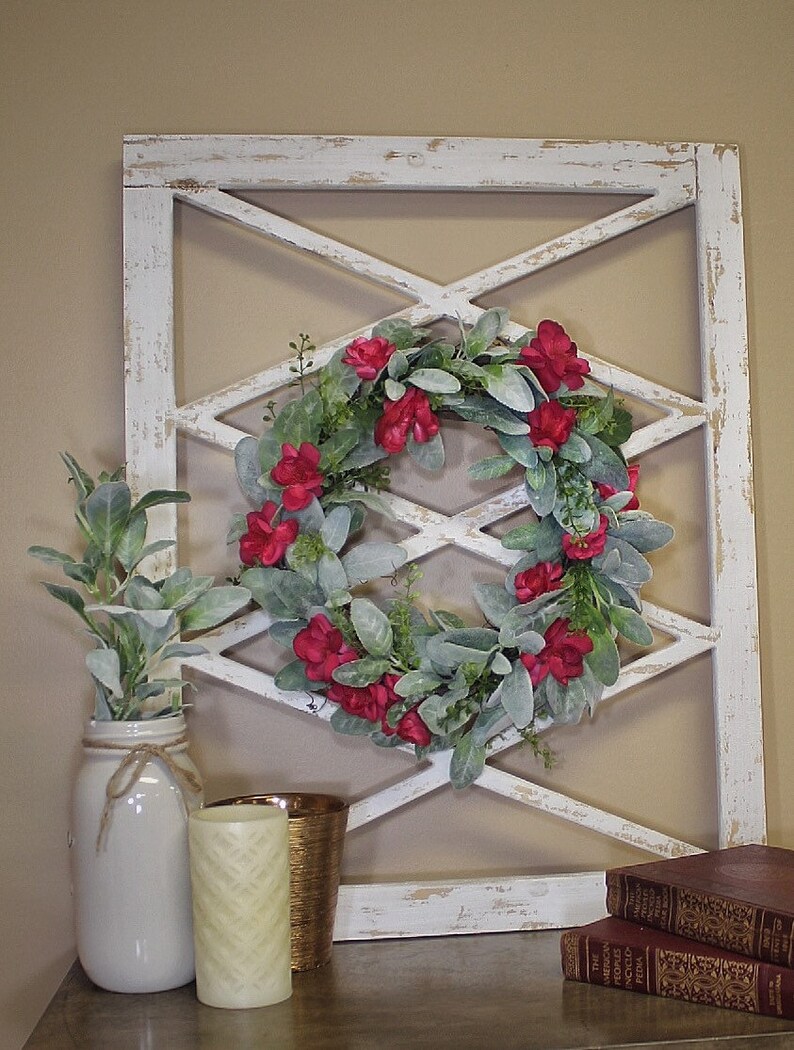 May include: A decorative white wooden wall hanging with a distressed finish, featuring a diamond-shaped lattice design. A wreath of red flowers and green leaves is centered within the lattice. A white jar with greenery, a candle, and books complete the display.