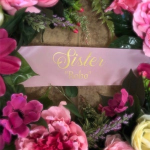 Puede incluir: Una corona floral rosa con una cinta rosa que dice "Sister "Bobo"" en letras doradas.