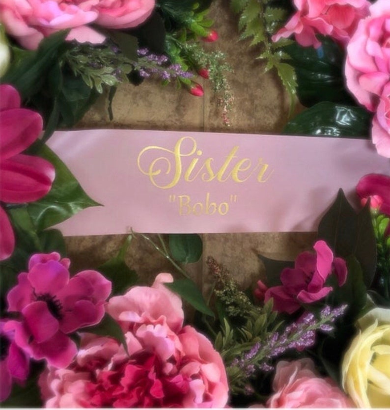 Puede incluir: Una corona floral rosa con una cinta rosa que dice "Sister "Bobo"" en letras doradas.