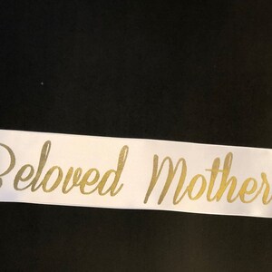 Puede incluir: Cinta de sat&eacute;n blanca con las palabras "Beloved Mother" escritas en letras doradas cursivas.