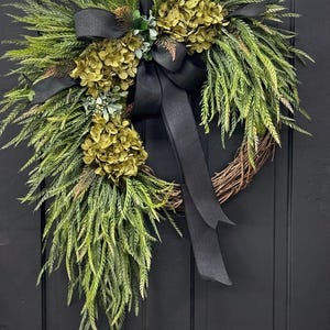 May include: A green and black wreath with a black ribbon bow. The wreath is made of greenery and has a few green hydrangea blooms.