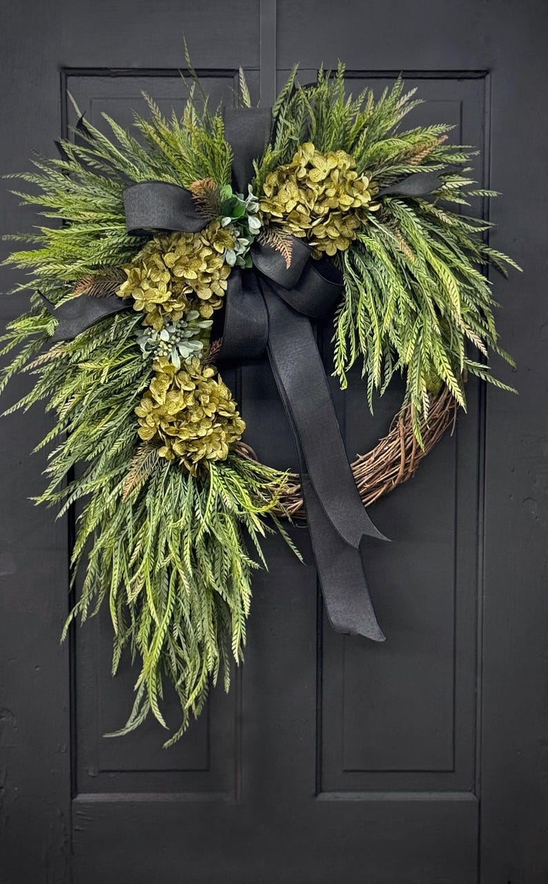 May include: A green and black wreath with a black ribbon bow. The wreath is made of greenery and has a few green hydrangea blooms.
