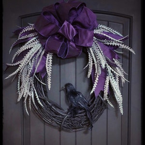 May include: A purple and white Halloween wreath with a black raven perched on the base. The wreath is made of grapevine and has a large purple bow.