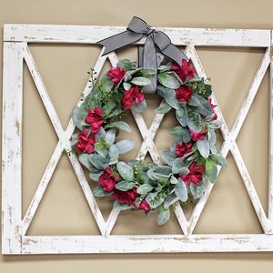 May include: A white, distressed wooden wall decor piece with a diamond-shaped lattice design. A wreath of red flowers and green leaves is centered within the diamond. A black and white striped bow is at the top.