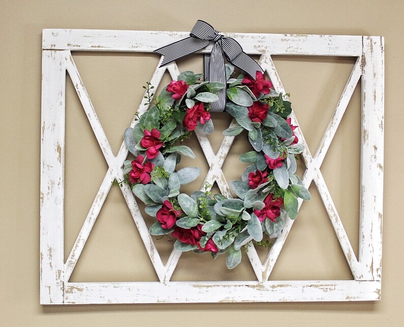 May include: A white, distressed wooden wall decor piece with a diamond-shaped lattice design. A wreath of red flowers and green leaves is centered within the diamond. A black and white striped bow is at the top.