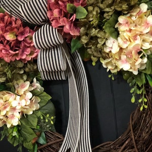 Pu&ograve; includere: Una ghirlanda fatta di ramoscelli marroni con fiori di ortensia rosa e bianchi e foglie verdi. Un nastro a righe bianche e nere &egrave; legato in un fiocco in cima alla ghirlanda.