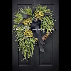 Puede incluir: Una corona decorativa con follaje verde, hortensias verde oliva y una cinta negra. La corona está hecha de ramitas tejidas y se exhibe en una puerta negra. Copyright 2025FIVESTARFLORAL.INC está impreso en la imagen.