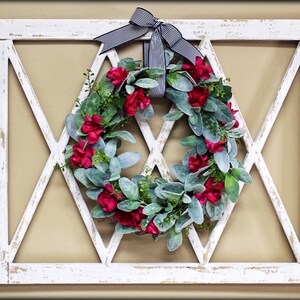 May include: A white, distressed-look wooden wall decor piece with a diamond-patterned design. A wreath of red flowers and green leaves is centered within the frame, and a black and white striped bow is at the top.
