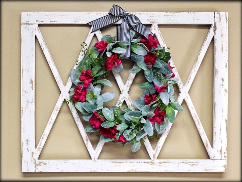 May include: A white, distressed-look wooden wall decor piece with a diamond-patterned design. A wreath of red flowers and green leaves is centered within the frame, and a black and white striped bow is at the top.