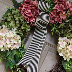 Pu&ograve; includere: Una ghirlanda di vite con ortensie rosa, bianche e verdi. Un nastro a righe bianche e nere &egrave; legato in un fiocco al centro della ghirlanda.