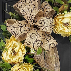 Pode incluir: Uma coroa de juta com um grande la&ccedil;o marrom com um padr&atilde;o de espiral. A coroa &eacute; decorada com flores de seda amarelas e folhas verdes.