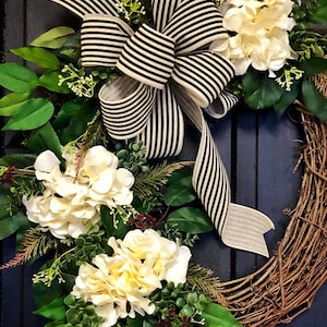 Puede incluir: Una corona de vid con flores de hortensia blancas y vegetación. Una cinta a rayas blancas y negras está atada en un lazo en la corona.