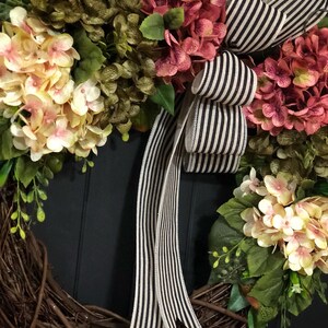 Pu&ograve; includere: Una ghirlanda fatta di ramoscelli marroni con fiori di ortensia rosa e crema e foglie verdi. Un nastro a righe bianche e nere &egrave; legato in un fiocco al centro.