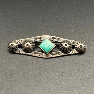 以下が含まれることがあります： 中央にターコイズ色の正方形の石をあしらったシルバーカラーのブローチ。ブローチは、花柄と矢印のデザインが施された長方形の形をしています。ブローチの長さは約7.6cmです。