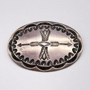 以下が含まれることがあります： 複雑なデザインの楕円形の銀色の金属バックル。中央には、ダイヤモンドの形を交差する2本の矢があり、詳細なパターンとスカラップエッジで囲まれています。バックルはおそらくヴィンテージです。