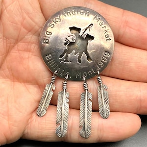 May include: A silver-coloured round pendant from Big Sky Indian Market, Billings, Mont. 1989, featuring a cutout of a person on horseback. The pendant has four dangling feather charms.