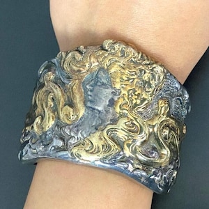 May include: A wide, ornate cuff bracelet with a silver and gold finish. The bracelet features a detailed relief of a classical female figure with flowing hair and intricate swirling patterns. The metalwork has a tarnished, antique appearance.